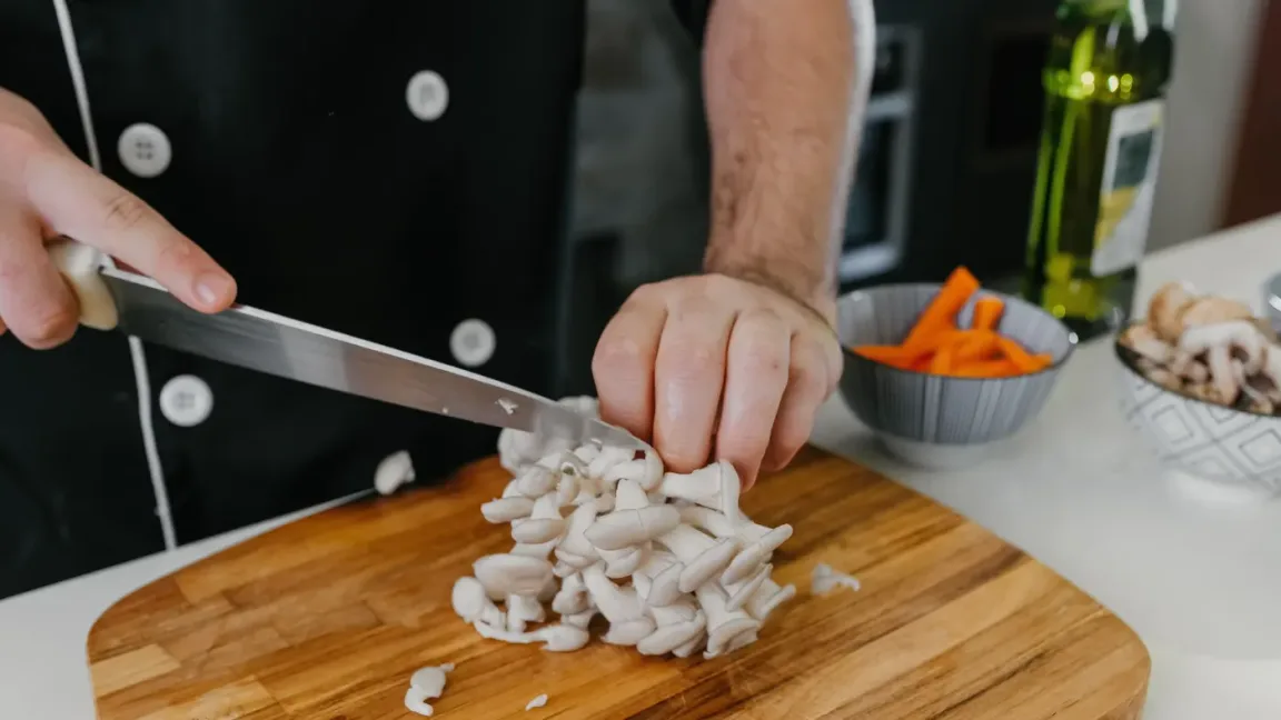 Eine Person in schwarzer Kochkleidung schneidet mit einem Messer Austernpilze auf einem Holzbrett; daneben stehen Schalen mit Möhren und weiteren Pilzen sowie eine Flasche Öl.