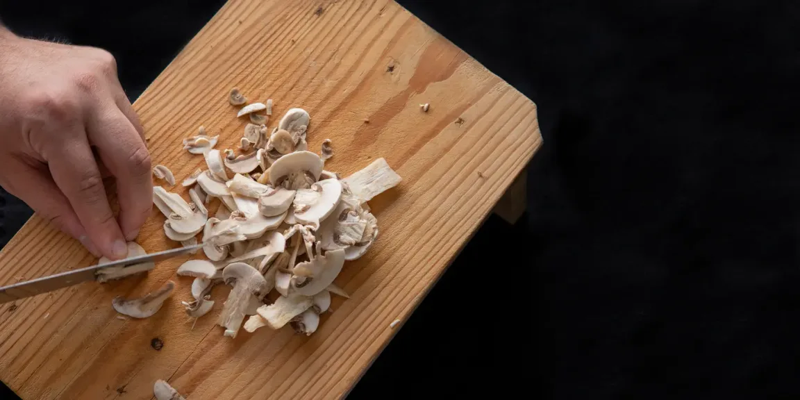 Eine Hand schneidet mit einem Messer weiße Champignons auf einem hölzernen Schneidebrett, auf dem bereits viele Pilzscheiben und -stücke liegen.
