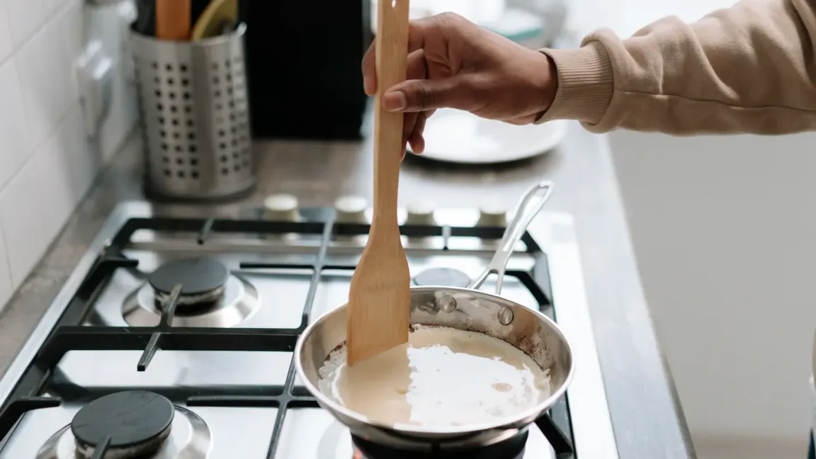 Eine Person rührt mit einem Holzlöffel in einer hellen, cremigen Sauce in einem Topf auf einem Gasherd; im Hintergrund stehen Küchenutensilien.