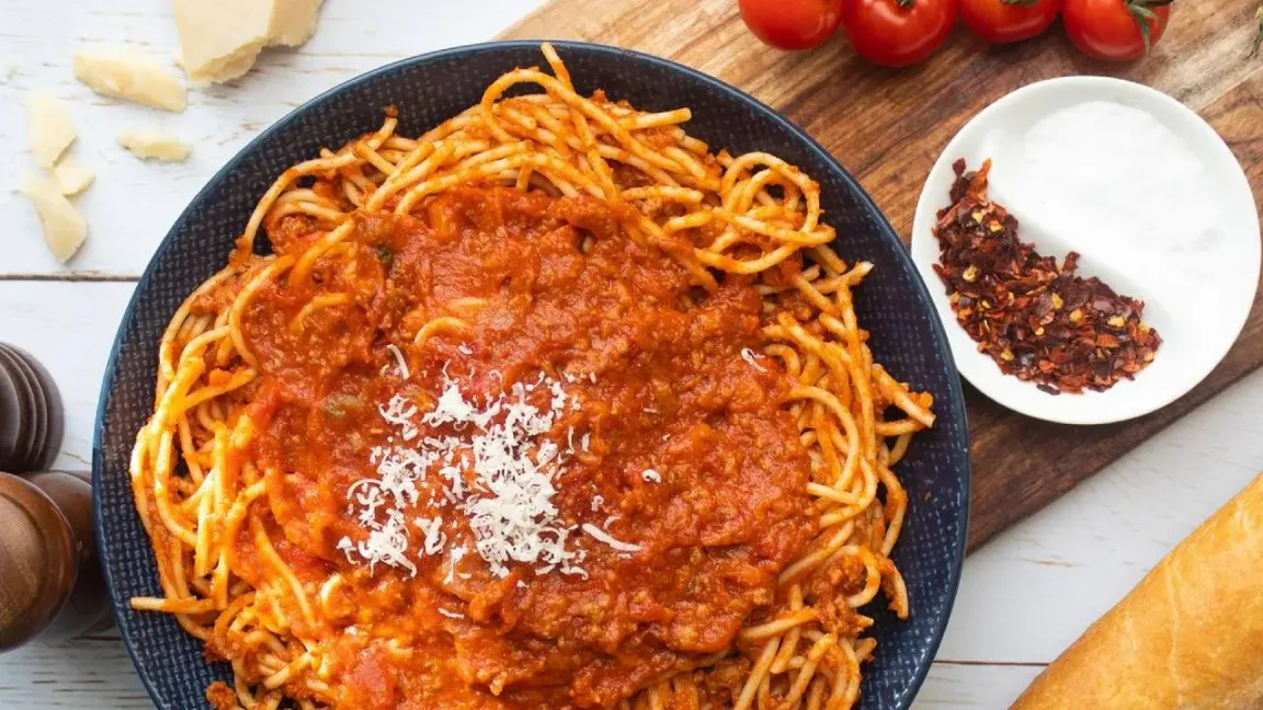 Auf einem dunklen Teller liegen Spaghetti mit roter, hackfleischhaltiger Tomatensauce und geriebenem Käse; daneben stehen Schälchen mit Salz und Chiliflocken, außerdem sind Tomaten und Brot auf einer Küchenarbeitsfläche zu sehen.