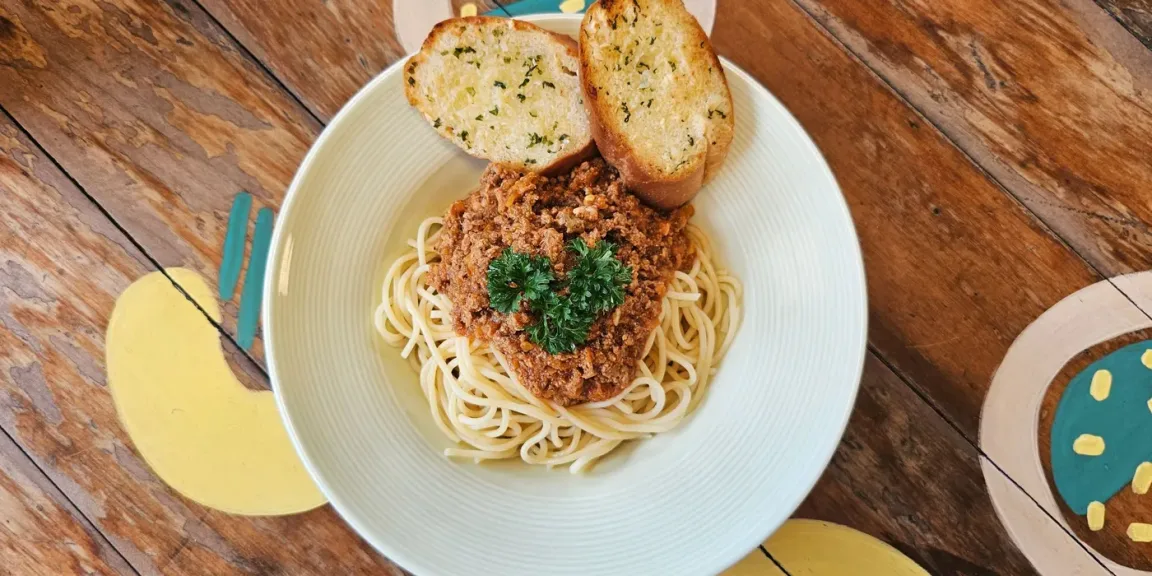 Auf einem Teller liegen Spaghetti mit einer Hackfleisch-Tomatensauce, garniert mit Petersilie, daneben liegen zwei Scheiben Knoblauchbrot auf einem Holztisch.