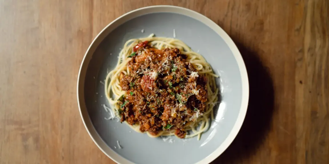 Auf einem grauen Teller liegt auf einem Holztisch ein Teller Spaghetti mit einer Hackfleisch-Tomatensauce, geriebenem Käse und Kräutern.