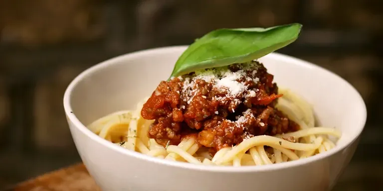 Eine Schüssel mit Spaghetti, darauf eine stückige rote Hackfleisch-Tomatensauce, geriebener Käse und ein Basilikumblatt, steht auf einer hölzernen Küchenfläche.