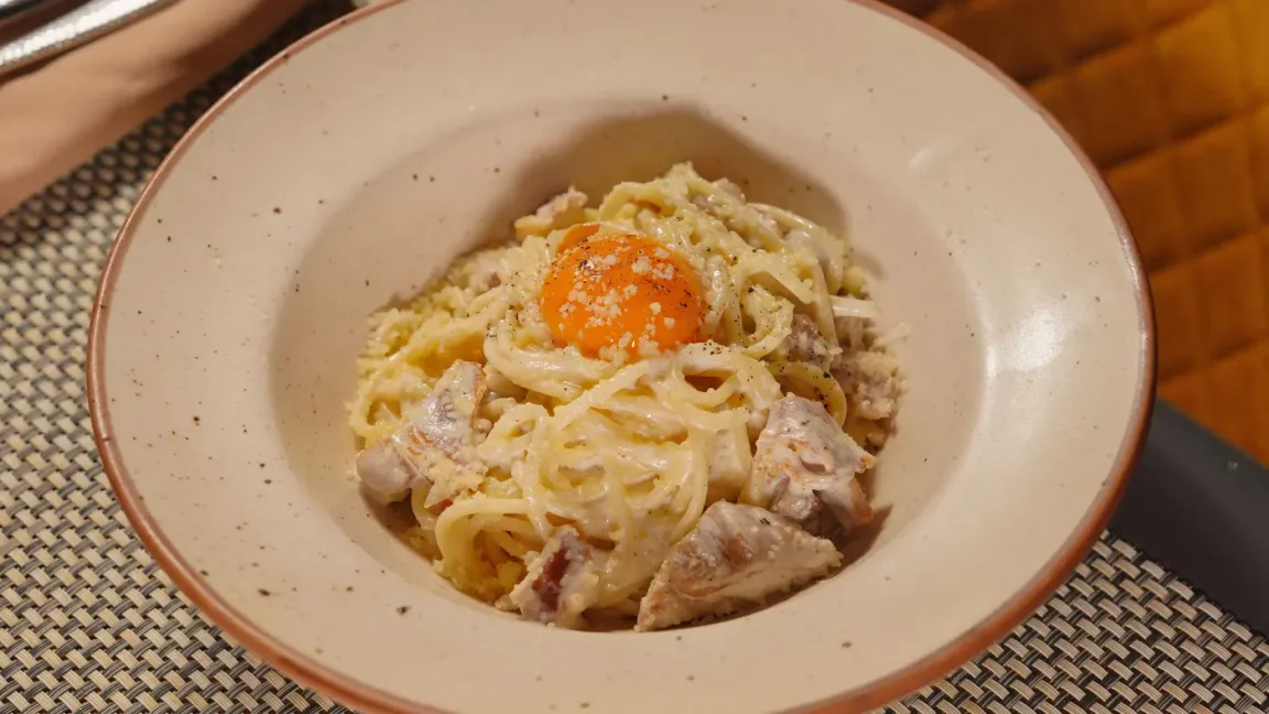 In einer hellen Keramikschüssel liegt ein Teller Spaghetti mit cremiger Sauce, Speck- oder Fleischstücken, geriebenem Käse, Pfeffer und einem rohen Eigelb in der Mitte auf einem Tischset.