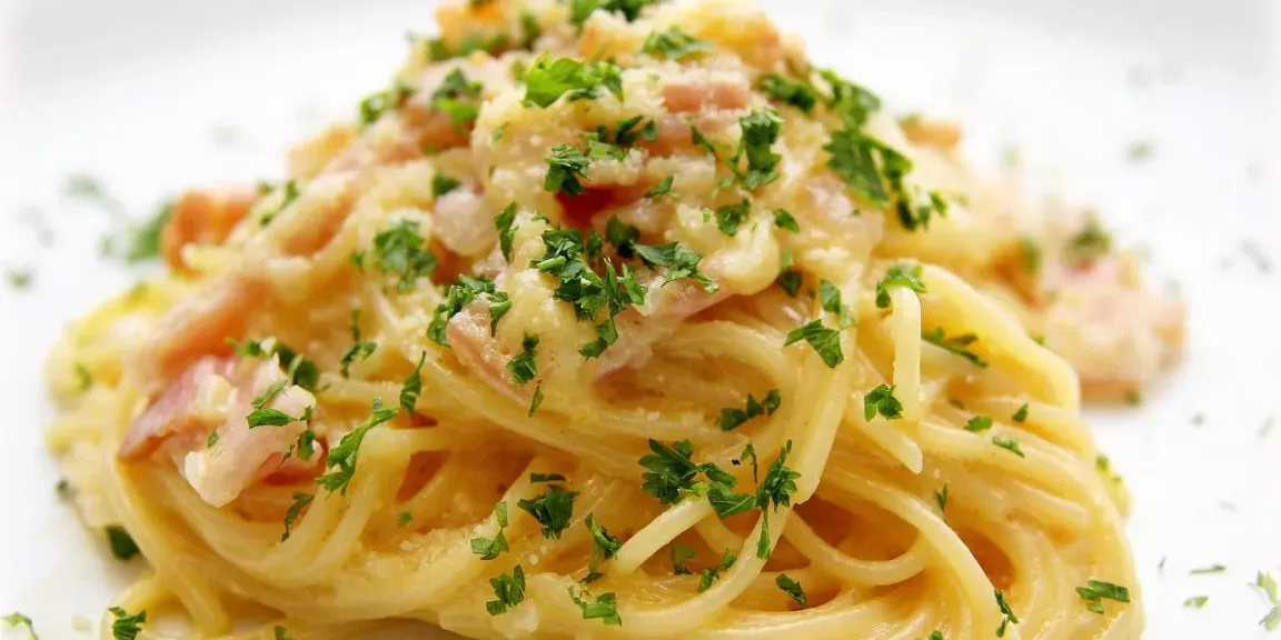 Auf einem weißen Teller liegt ein Berg Spaghetti mit cremiger heller Sauce, Speckstücken und gehackter grüner Petersilie in heller Küchenumgebung.