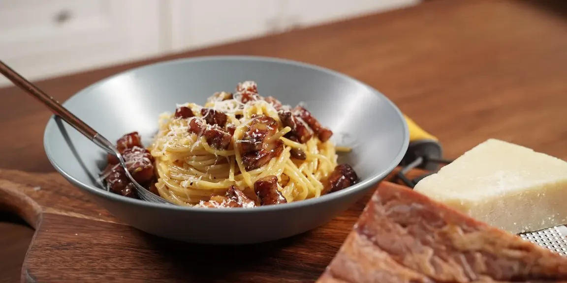 Auf einem Holztisch steht eine graue Schale mit Spaghetti Carbonara und knusprigen Speckstücken, daneben liegen ein Stück Hartkäse und eine Reibe, während eine Gabel in der Pasta steckt.