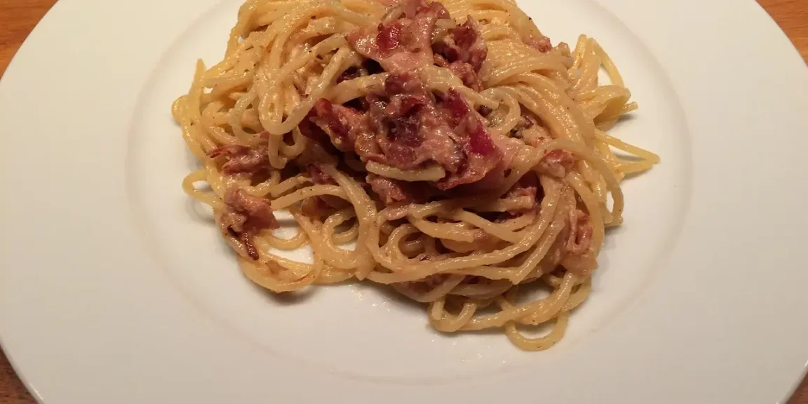 Auf einem weißen Teller liegt ein Tellergericht aus Spaghetti mit cremiger heller Sauce und gebratenen Speckstücken auf einer Holzoberfläche.