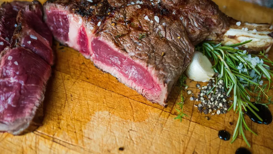 Auf einem hölzernen Schneidebrett liegen ein angeschnittenes, innen rosarotes Steak und mehrere Fleischscheiben, daneben befinden sich Rosmarinzweige, Knoblauch, grobes Salz, Pfeffer und dunkle Soße.