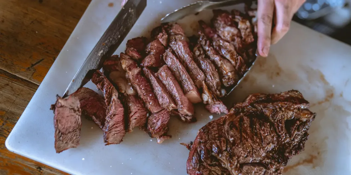 Auf einem weißen Schneidebrett liegen ein gebratenes Steak und mehrere aufgeschnittene Fleischstücke, während eine Person sie mit einem Messer und einer Zange bearbeitet.