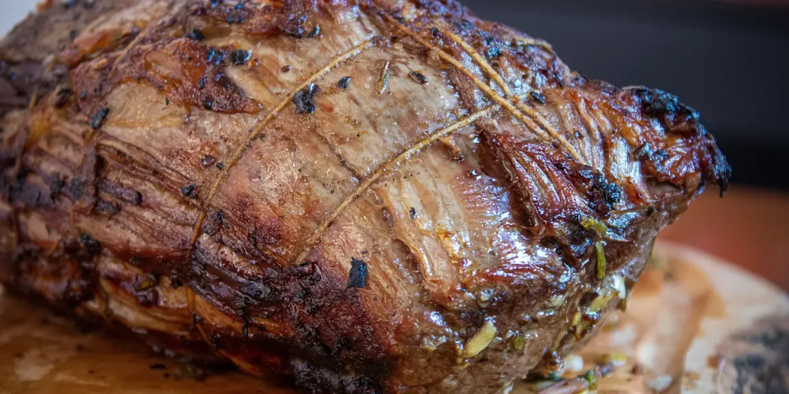 Auf einem hölzernen Schneidebrett liegt ein großer gebratener, mit Küchengarn verschnürter Fleischbraten, dessen Oberfläche gebräunt und mit Kräutern gewürzt ist.