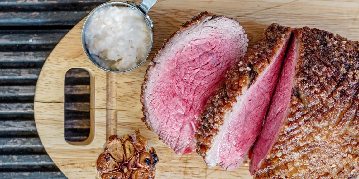 Auf einem Holzbrett liegen mehrere Scheiben eines rosa gebratenen Rinderbratens mit kräftiger Kruste, daneben eine kleine Schale mit heller Beilage und eine halbierte geröstete Knoblauchknolle.