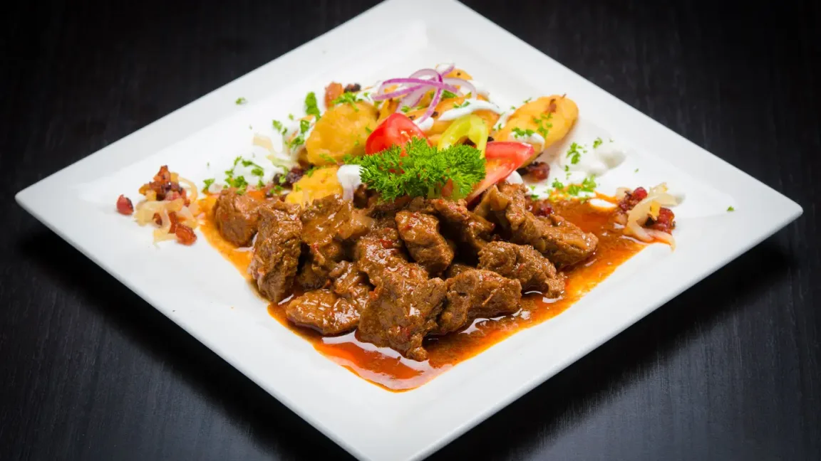 Auf einem weißen quadratischen Teller liegt Gulasch in brauner Soße, garniert mit Petersilie und Gemüse sowie Beilagen, auf einem dunklen Tisch.
