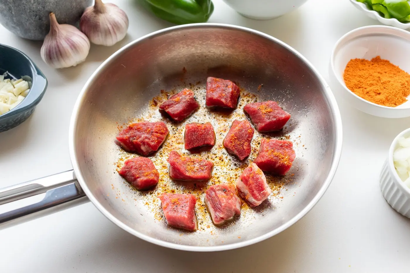 Rohe gewürzte Rinderwürfel in einer Pfanne, daneben Knoblauch und Paprikapulver