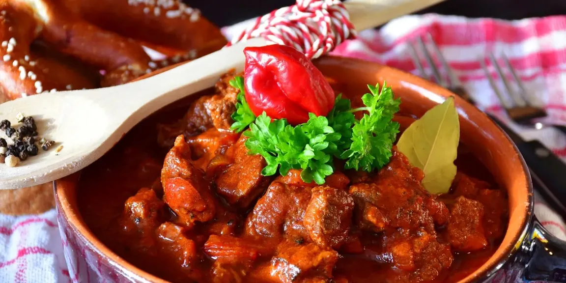 Zu sehen ist eine Schale mit rotbraunem Gulasch aus Fleischstücken und Gemüse, garniert mit Petersilie und einem Lorbeerblatt, daneben liegen ein Holzlöffel mit Pfefferkörnern und weitere Küchenutensilien auf einem Tuch.
