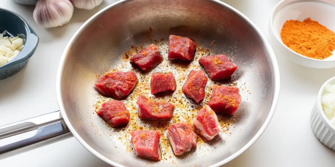 In einer großen Metallpfanne braten mehrere gewürzte Stücke rohen roten Fleisches, daneben stehen Schalen mit gehacktem Knoblauch, Zwiebeln und Gewürzpulver sowie ganze Knoblauchknollen auf einer hellen Küchenarbeitsfläche.