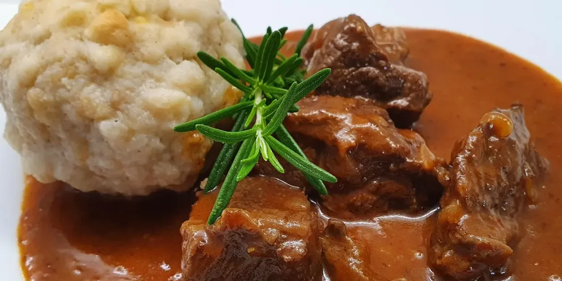 Auf einem weißen Teller liegen mehrere Stücke Gulasch in brauner Soße neben einem großen Semmelknödel, garniert mit einem Rosmarinzweig.