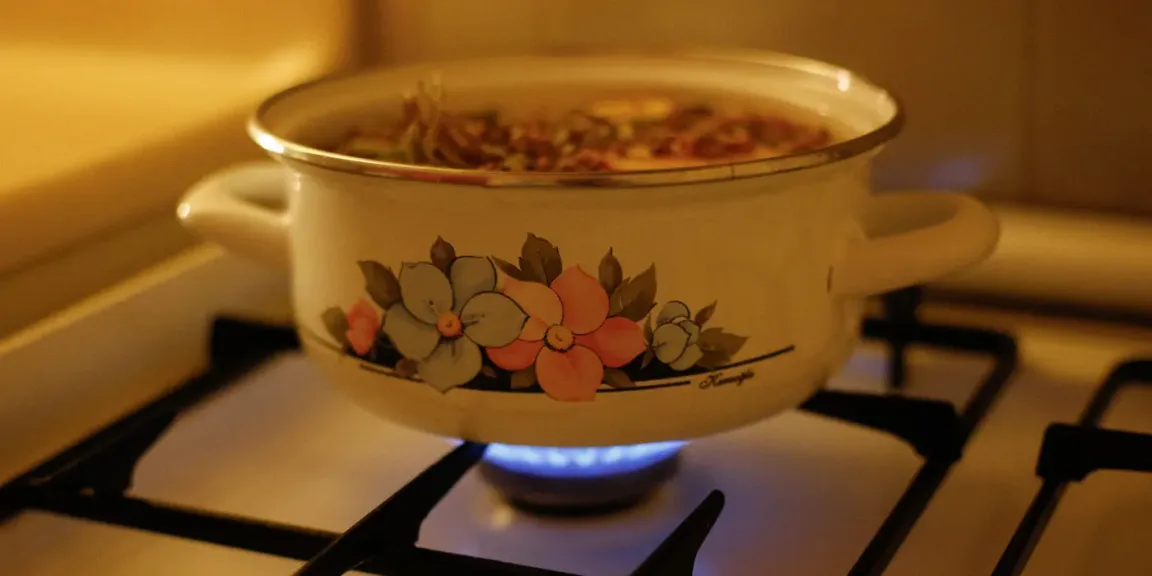 Ein emaillierter, floral verzierter Topf mit einem sichtbaren Gulasch- oder Eintopfgericht steht auf einer eingeschalteten Gasflamme auf einem Herd.