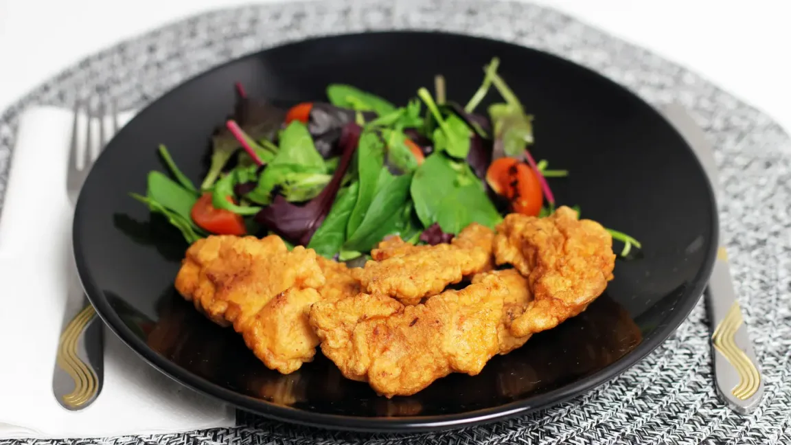 Auf einem schwarzen Teller liegen mehrere goldbraun panierte Hähncheninnenfilets neben einem gemischten Blattsalat mit Tomaten, auf einem gedeckten Tisch.