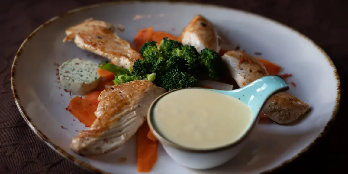 Auf einem großen Teller liegen mehrere gebratene Hähncheninnenfilets mit Brokkoli und Karotten, daneben stehen eine Schale mit heller Soße und ein Klecks Kräuterbutter.