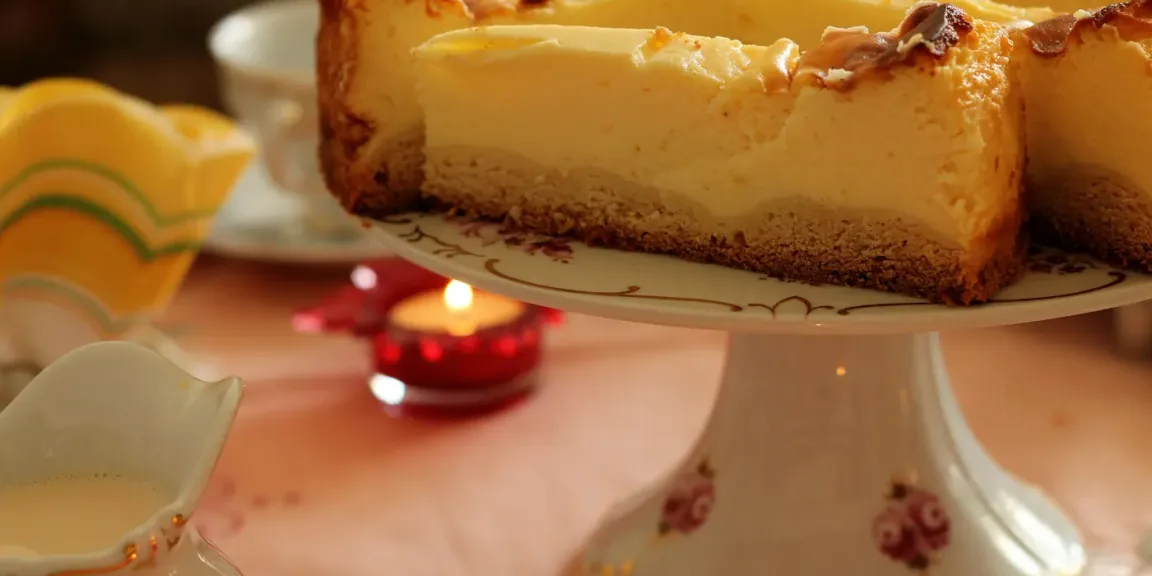 Auf einer dekorativen Tortenplatte steht ein angeschnittener Käsekuchen mit gebräunter Oberfläche und Keksboden, daneben stehen ein Kännchen und eine brennende Kerze auf einem gedeckten Tisch.
