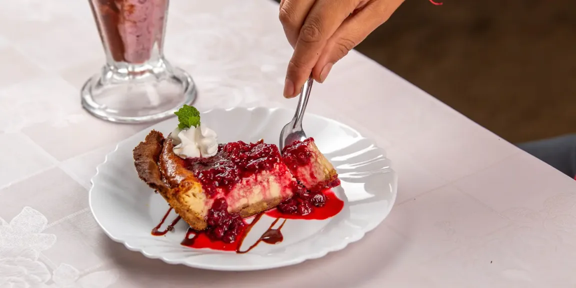Auf einem weißen Teller liegt ein Stück Käsekuchen mit roter Beerensoße, Schlagsahne und Minze, während eine Hand mit einer Gabel ein Stück davon nimmt; daneben steht ein Glas mit einem Getränk auf einem gedeckten Tisch.