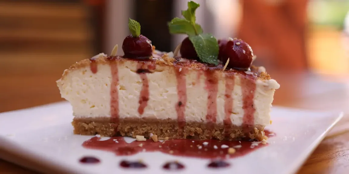 Auf einem weißen Teller steht ein Stück Käsekuchen mit Keksboden, roter Fruchtsauce, Kirschen und Minzblättern auf einem Holztisch.