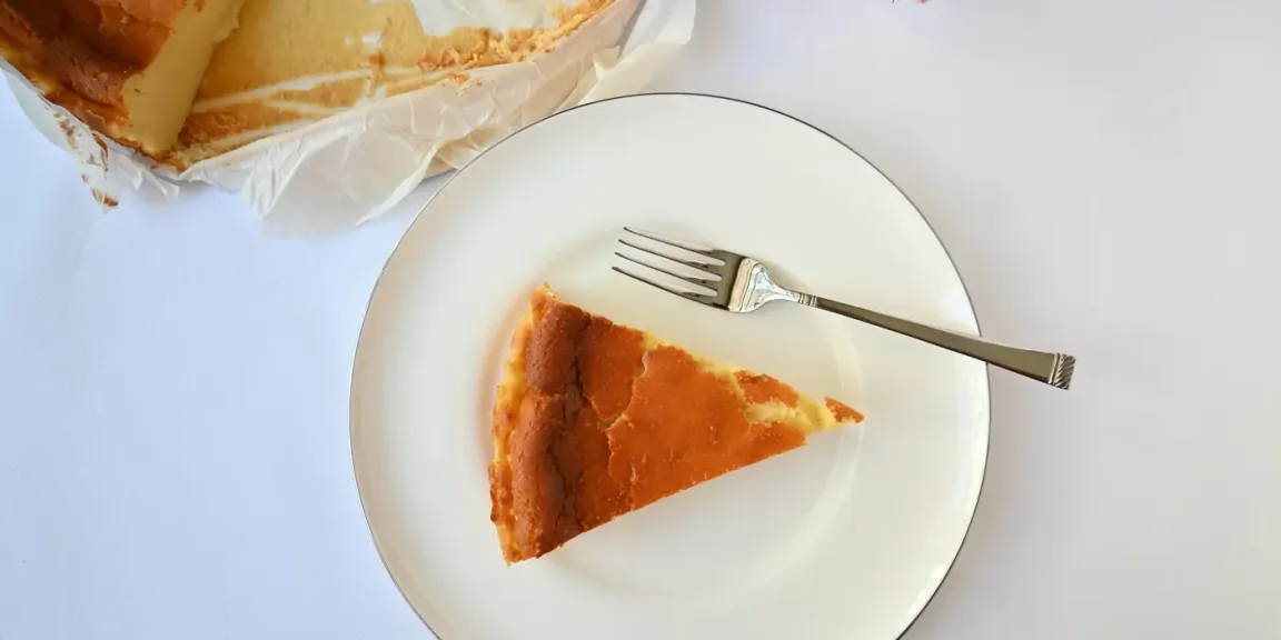 Auf einem weißen Teller liegt ein Stück Käsekuchen mit deutlich gebräunter, rissiger Oberfläche und daneben eine Gabel; im Hintergrund ist ein weiterer Teil des Kuchens auf Backpapier zu sehen.