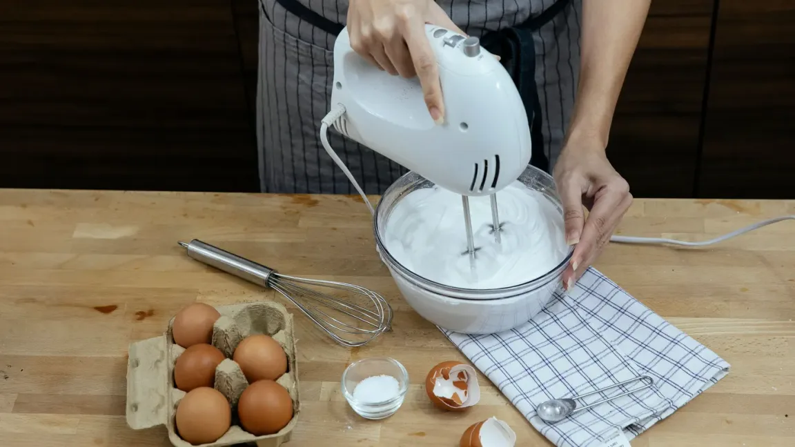 Eine Person schlägt in einer Glasschüssel mit einem elektrischen Handmixer Eiweiß zu Schaum, daneben liegen Eier, ein Schneebesen, eine Schale mit Zucker und zerbrochene Eierschalen auf einer Küchenarbeitsplatte.
