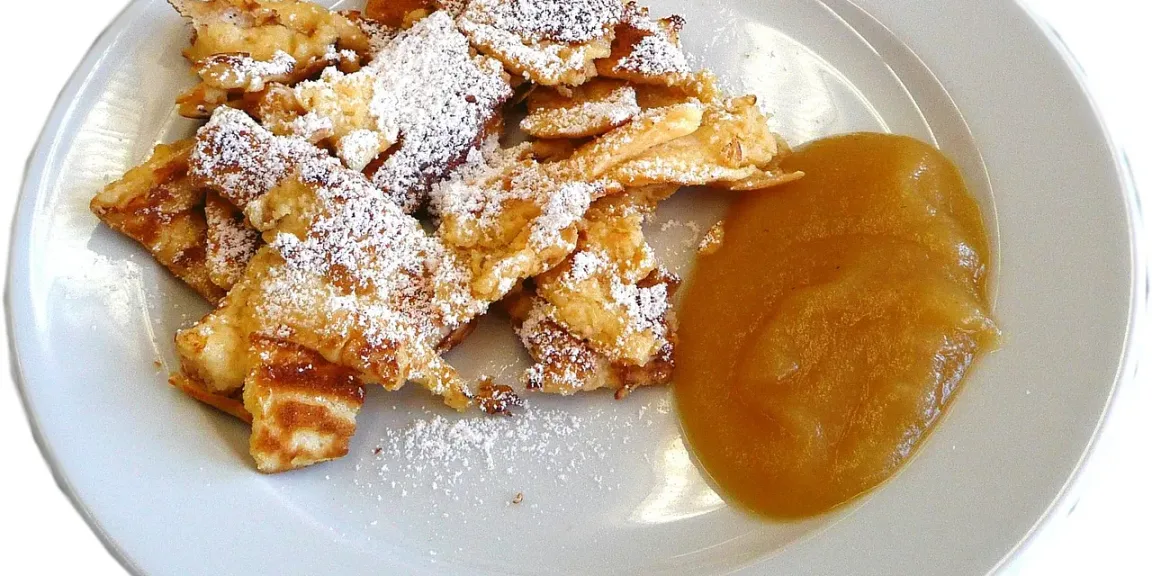 Auf einem weißen Teller liegen zerrissene, goldbraun gebratene Stücke Kaiserschmarrn mit Puderzucker und einem Klecks Apfelmus.