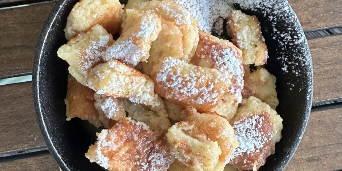 In einer schwarzen Pfanne liegen unregelmäßig zerrissene, goldbraun gebratene Kaiserschmarrnstücke, die mit Puderzucker bestäubt sind, auf einem Holztisch.