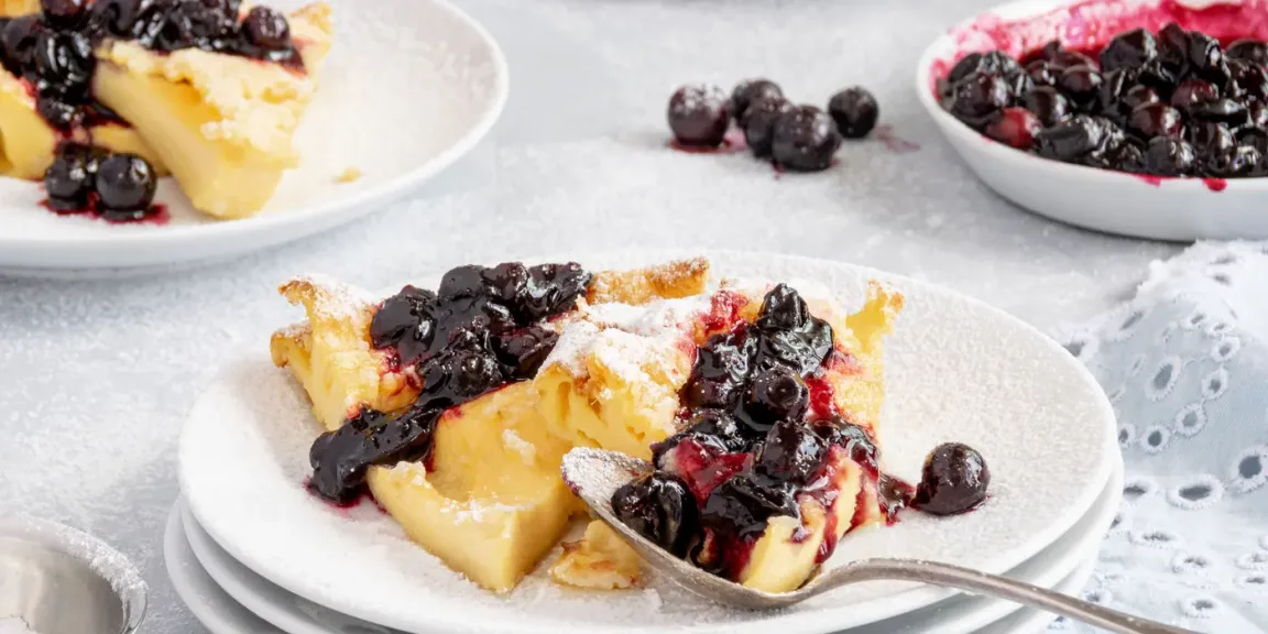 Auf einem weißen Teller liegen goldbraun gebratene Stücke Kaiserschmarrn mit Puderzucker und dunkler Beerensoße, daneben stehen Schalen mit Beeren und im Vordergrund liegt ein Löffel auf einer hell gedeckten Küchenfläche.
