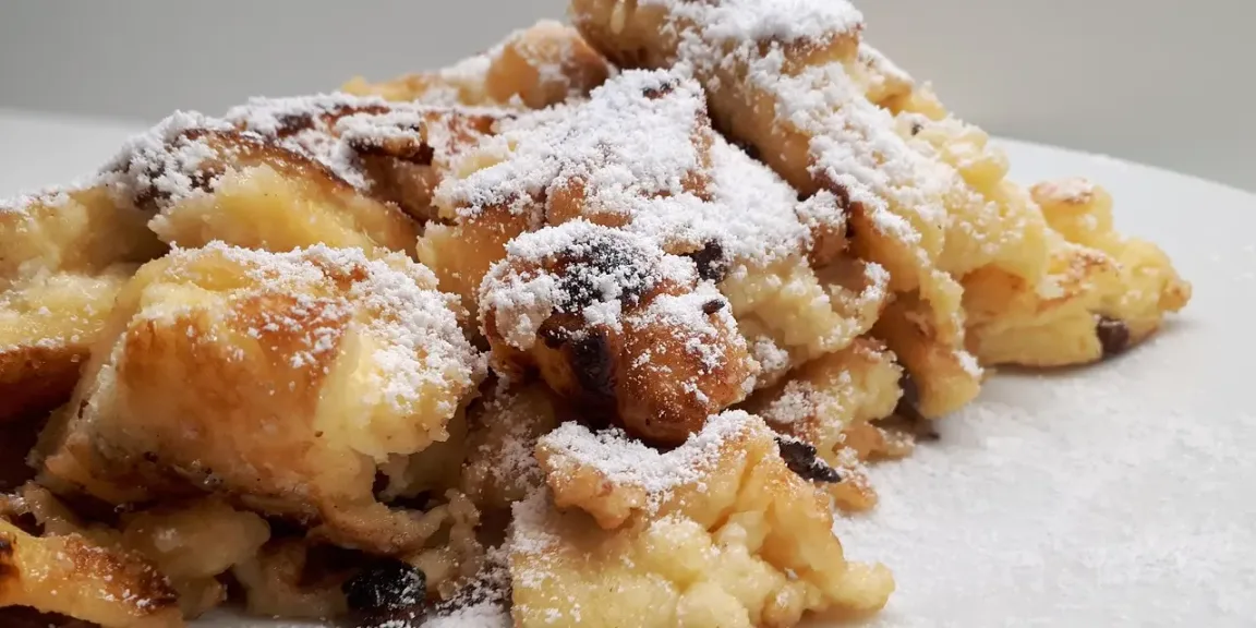 Auf einem weißen Teller liegt ein Haufen in Stücke gerissener, goldbraun gebratener Kaiserschmarrn mit Rosinen und einer dichten Schicht Puderzucker.