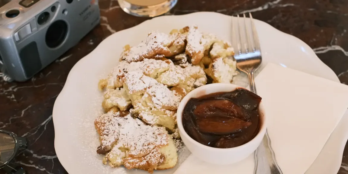 Auf einem weißen Teller liegt in Stücke gerissener, mit Puderzucker bestäubter Kaiserschmarrn neben einer Schale mit dunklem Kompott; daneben liegen eine Gabel und ein Messer auf einer Serviette, im Hintergrund steht eine Kamera.