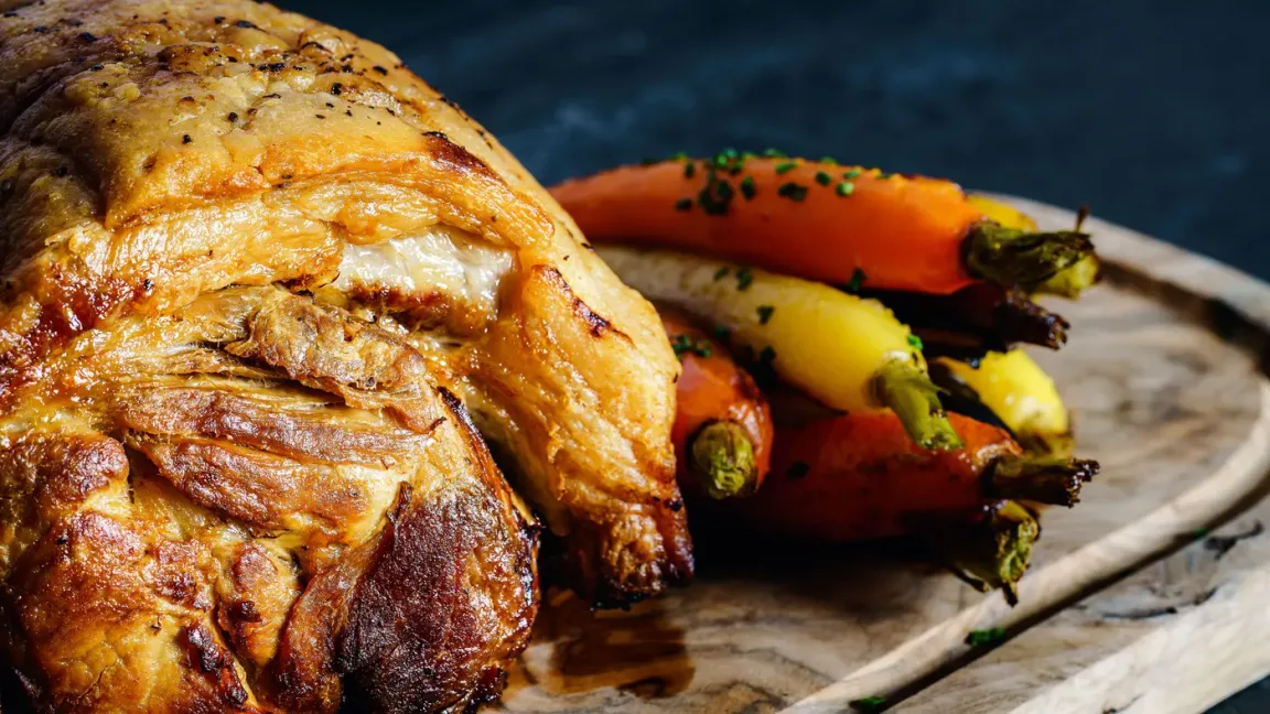 Auf einem Holzbrett liegt ein großer gebratener Schweinebraten mit knuspriger Kruste, daneben liegen geröstete Möhren und Kartoffeln.