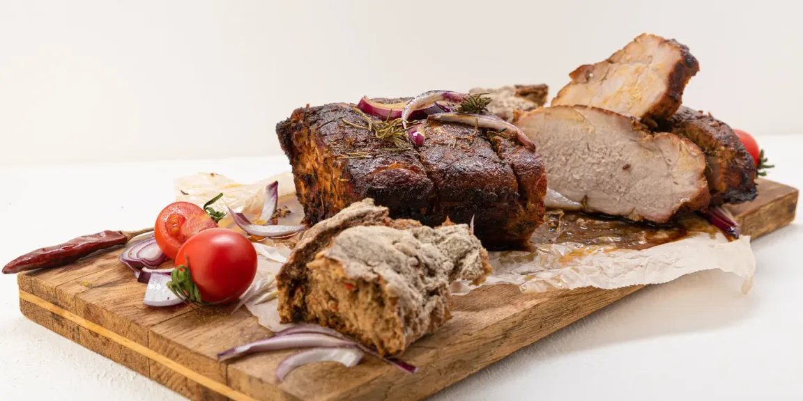 Auf einem Holzbrett liegen ein ungeschnittener und ein aufgeschnittener, dunkel gebratener Schweinebraten mit Kruste, dazu Brot, rote Zwiebeln, Tomaten und Kräuter.