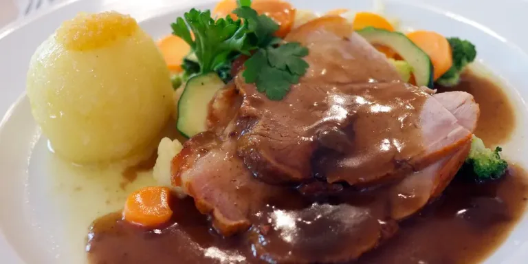 Auf einem weißen Teller liegen mehrere Scheiben eines gebratenen Schweinebratens mit dunkler Soße, einer Kartoffelklöße, Möhren, Zucchini, Brokkoli und Petersilie.