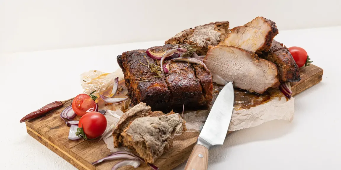 Auf zwei Holzschneidebrettern liegen mehrere Stücke aufgeschnittener Schweinebraten mit dunkler Kruste, dazu Zwiebeln, Tomaten, Knoblauch, Kräuter und ein Küchenmesser.