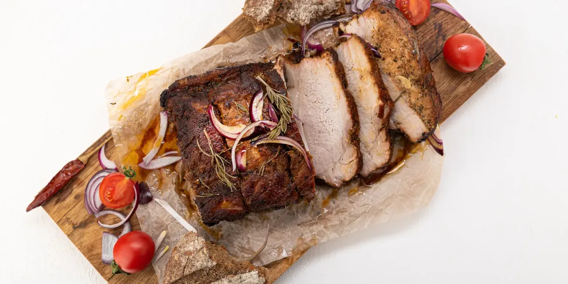 Auf einem hölzernen Schneidebrett liegt ein aufgeschnittener, gebräunter Schweinebraten auf Backpapier, garniert mit roten Zwiebeln, Rosmarin und Tomaten; daneben liegen Brotstücke und eine Chilischote.