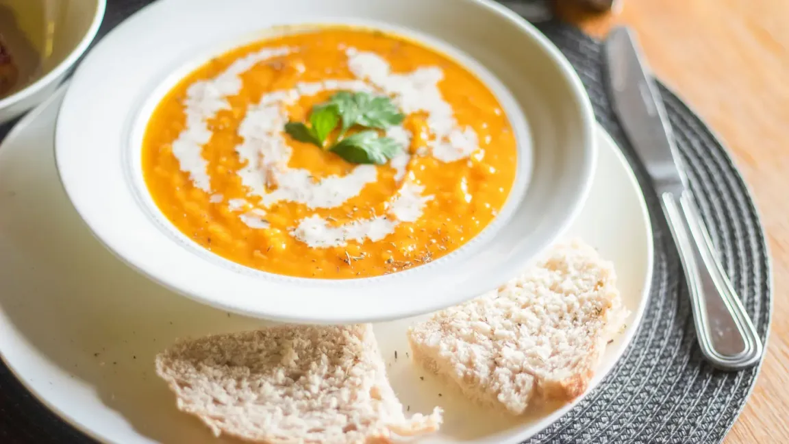 Auf einem weißen Teller steht eine orangefarbene Kürbissuppe mit einem Sahnewirbel und Petersilie, daneben liegen zwei Stücke Brot; Besteck liegt auf einer gewebten Tischunterlage.