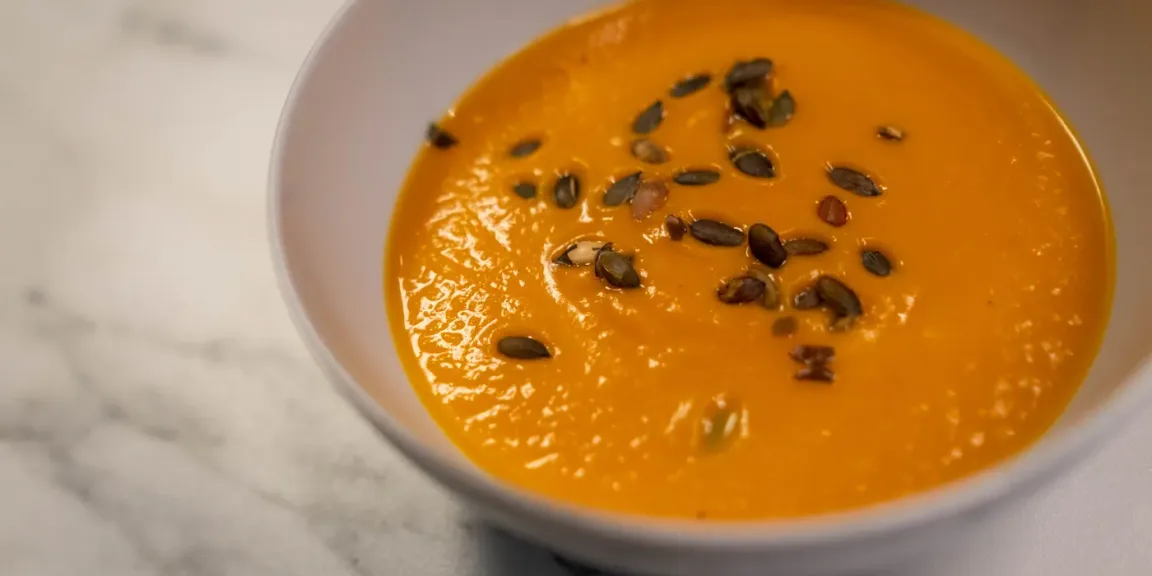 Eine Schale mit glatter orangefarbener Kürbissuppe steht auf einer hellen Küchenoberfläche und ist mit gerösteten Kürbiskernen bestreut.