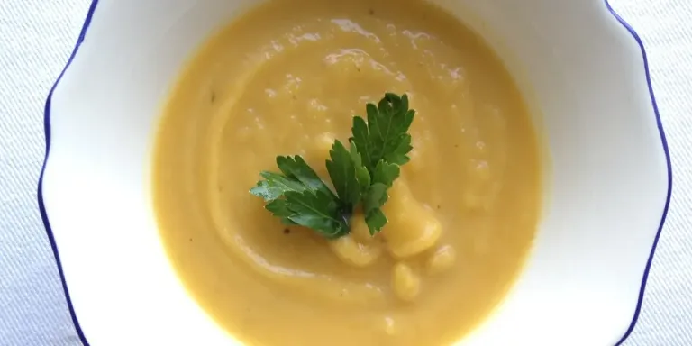 Eine Schale mit orangegelber, pürierter Kürbissuppe steht auf einem hellen Tuch und ist mit einem Zweig Petersilie garniert.