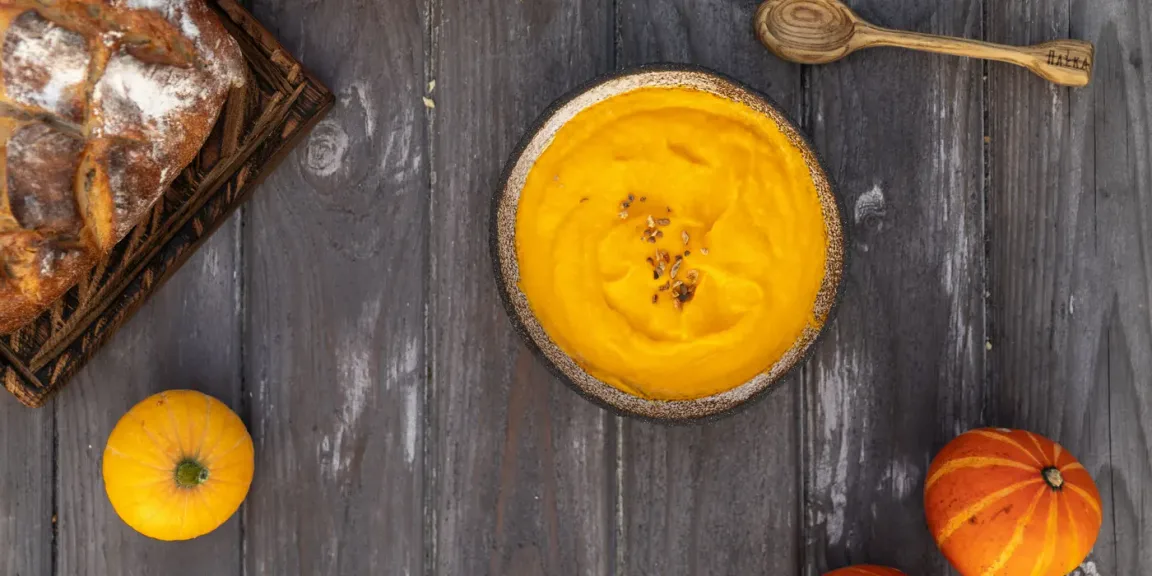 Auf einem dunklen Holztisch stehen eine Schale mit orangefarbener Kürbissuppe, mehrere Kürbisse, ein Brotlaib in einem Korb und ein Holzlöffel.