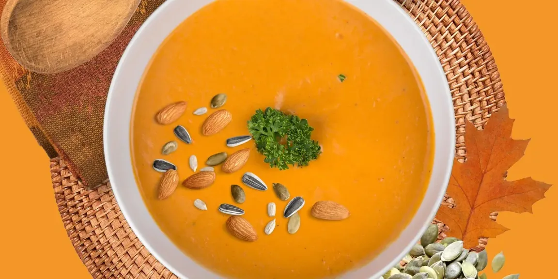 Eine Schale mit glatter orangefarbener Kürbissuppe steht auf einem geflochtenen Platzset und ist mit Petersilie, Mandeln und verschiedenen Kernen garniert; daneben liegen ein Holzlöffel und ein Herbstblatt.
