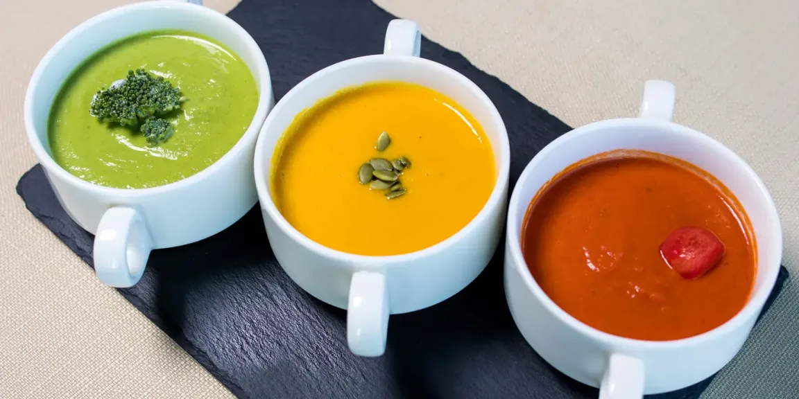 Auf einer schwarzen Schieferplatte stehen drei weiße Tassen mit pürierten Suppen: links eine grüne Suppe mit Brokkolistückchen, in der Mitte eine orange Kürbissuppe mit Kürbiskernen und rechts eine rote Suppe mit einem Stück Tomate.