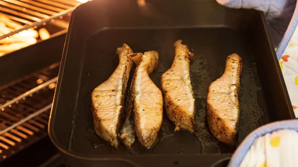 In einer dunklen Auflaufform liegen vier gebratene Fischstücke mit Haut und Gewürzen, die offenbar in einem geöffneten Ofen stehen; am rechten Bildrand ist ein Geschirrtuch zu sehen.
