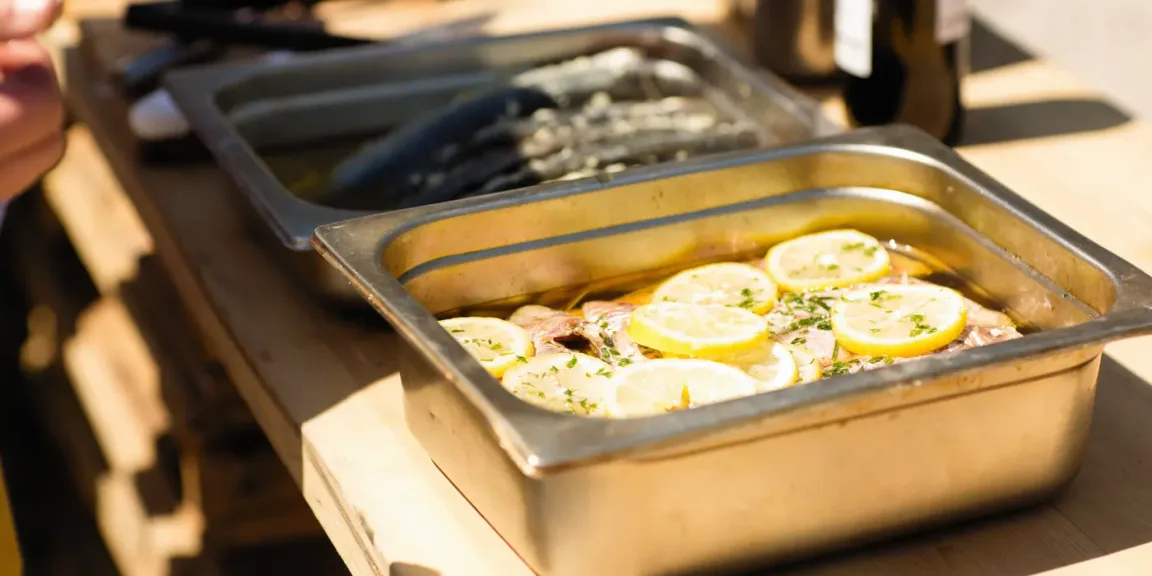In einer Edelstahl-Gastronomieschale liegen mehrere mit Zitronenscheiben und Kräutern belegte Fischfilets in Flüssigkeit, daneben stehen weitere Schalen in einer professionellen Küche.