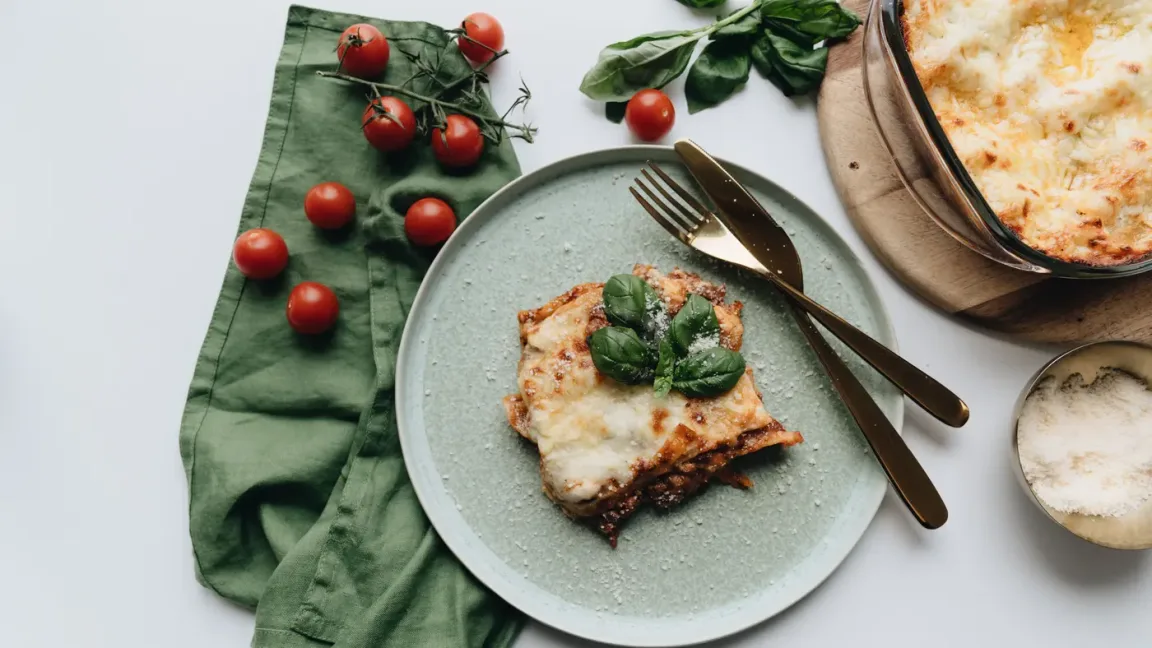 Auf einem Teller liegt ein Stück goldbraun überbackene Lasagne mit Basilikum, daneben stehen eine Auflaufform mit weiterer Lasagne, Tomaten, Basilikum, Besteck und eine Schale mit geriebenem Käse auf einer Küchenfläche.