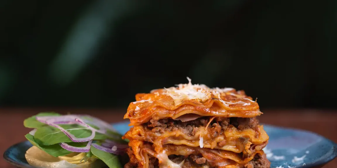 Auf einem blauen Teller steht ein Stück geschichtete Lasagne mit Hackfleisch, Tomatensauce und geriebenem Käse, daneben liegt ein kleiner Salat mit roten Zwiebeln und Dressing.