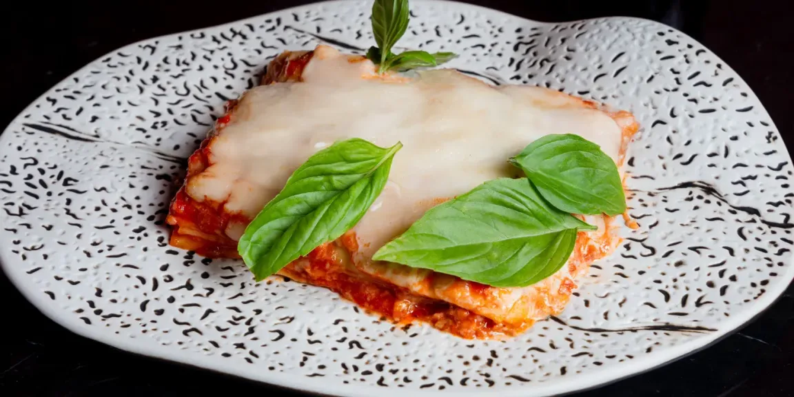 Auf einem gemusterten Teller liegt ein Stück Lasagne mit Tomatensauce, geschmolzenem Käse und frischen Basilikumblättern auf einer dunklen Oberfläche.