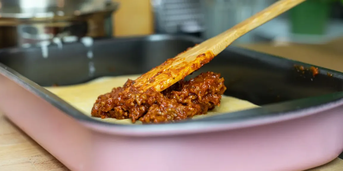 In einer rechteckigen Auflaufform wird mit einem Holzlöffel eine Schicht Hackfleischsoße auf helle Lasagneplatten gestrichen, während im Hintergrund eine Küche zu sehen ist.